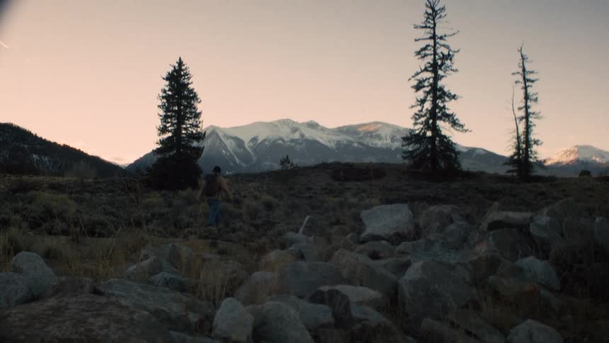 A hiker with a backpack walks through a rocky landscape towards snow-capped mountains at sunset