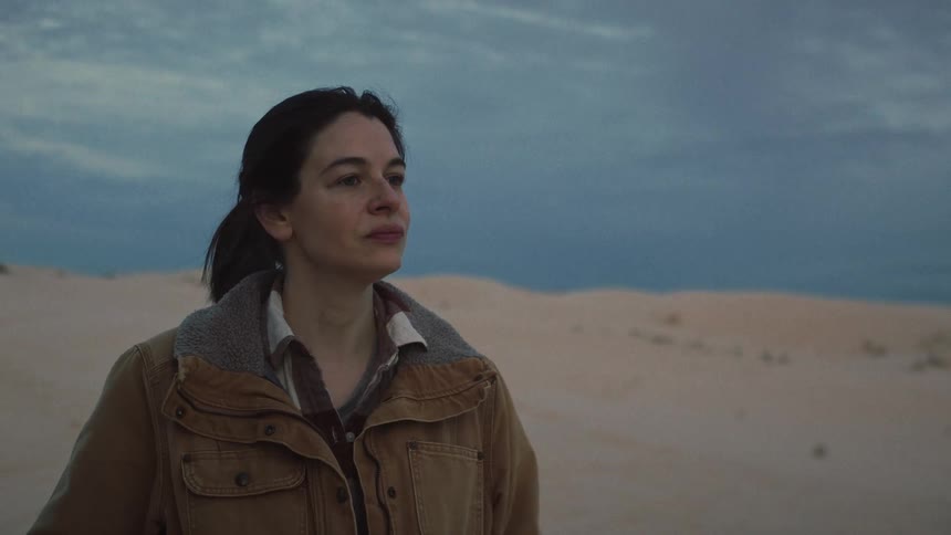 A thoughtful woman wearing a warm jacket stands alone in a vast desert landscape at dusk, looking off into the distance