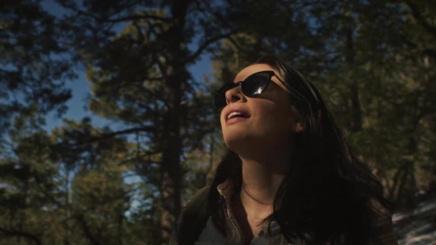 In slow motion, a smiling female hiker wearing sunglasses looks up through the trees, shielding her eyes and enjoying the warm sunlight