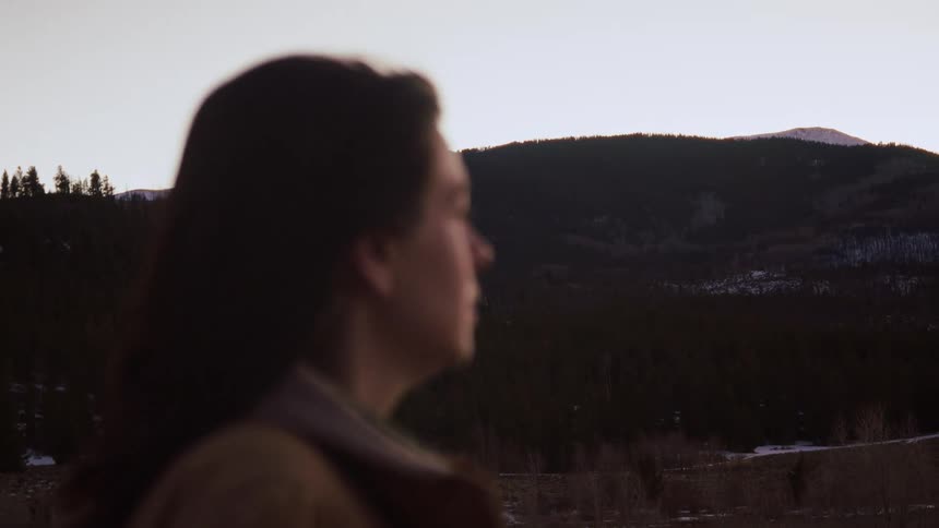 An out-of-focus person with long hair stands still, looking out at a vast, tree-covered mountain landscape at dusk