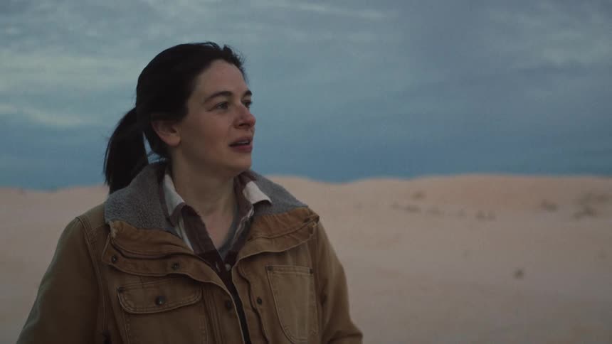 A thoughtful woman stands in a vast desert landscape at dusk, her expression a mixture of emotions as she reflects in solitude