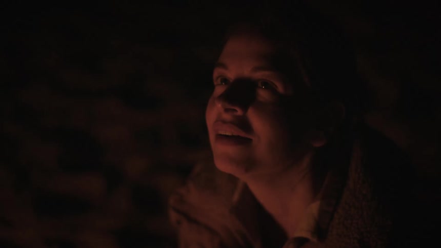 A woman's face is illuminated by a warm campfire at night as she smiles contentedly