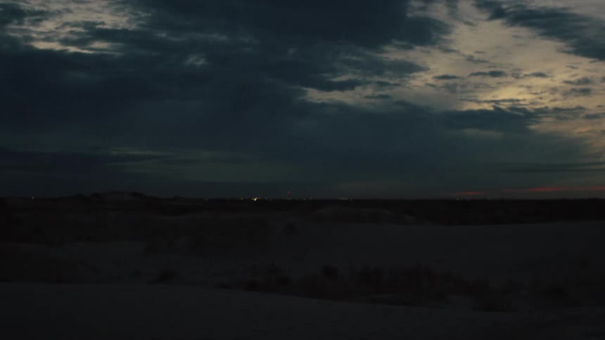 A dark desert landscape is silhouetted against a cloudy sky at dusk, with distant city lights twinkling on the horizon