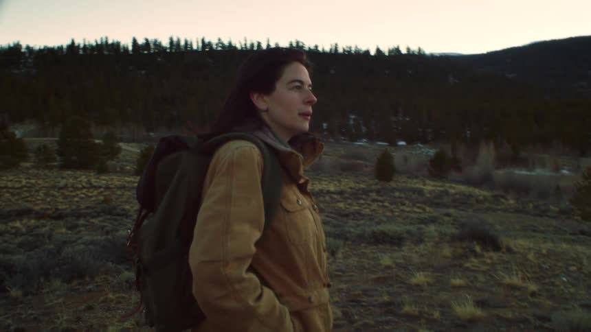 A solo female hiker with a backpack stops to admire the mountain view while trekking through a valley at dusk in slow motion
