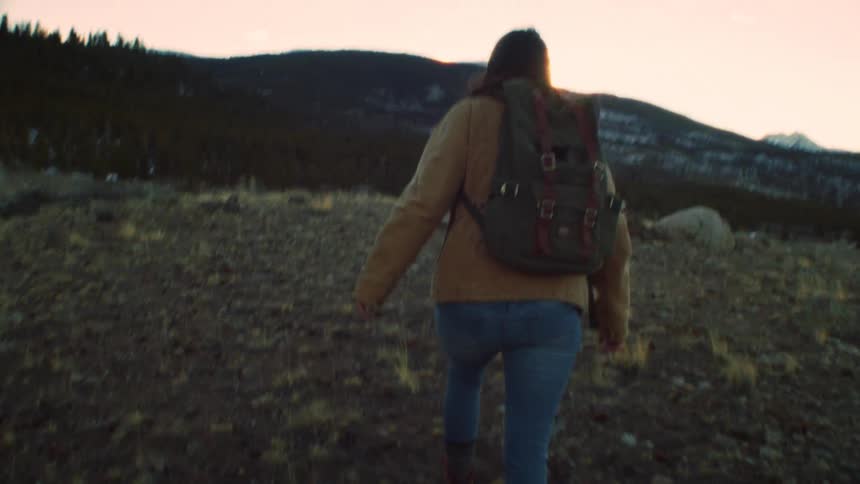 A woman with a backpack hikes up a rocky mountain trail towards the sunset