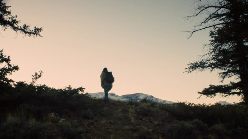A person with a backpack hikes up a hill toward a distant snow-capped mountain range at sunset