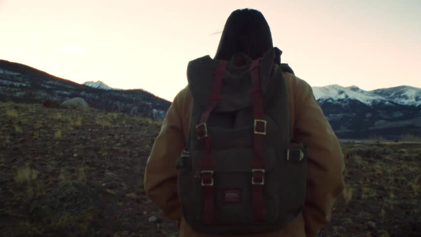A slow-motion tracking shot follows a woman with a backpack as she hikes through a scenic mountain landscape at dusk