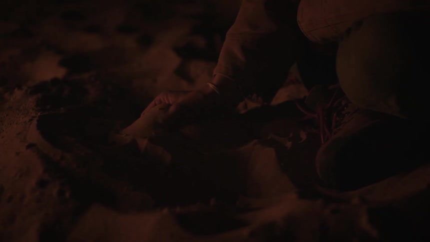A person's hand scoops up sand and lets it fall through their fingers in slow motion by the warm glow of a campfire at night