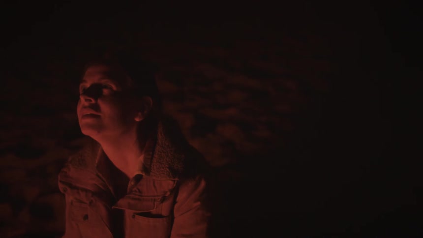 A young woman sits alone in the dark, her pensive face illuminated by the flickering red light of a campfire