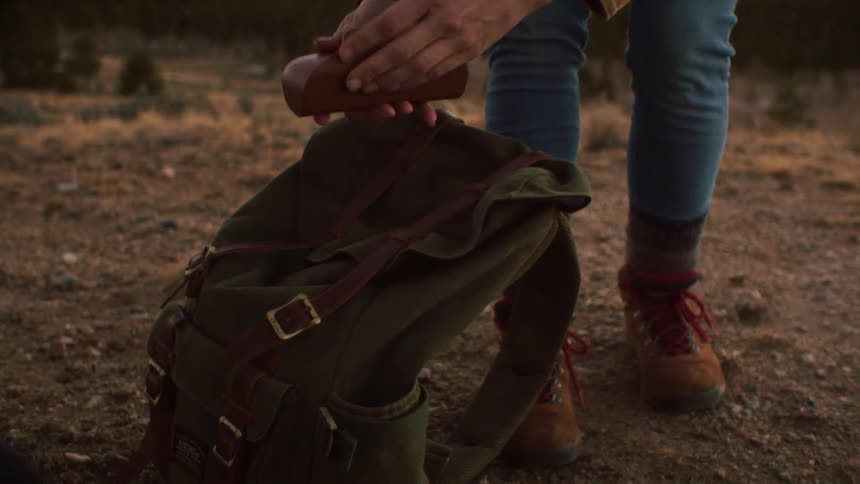 A hiker in boots takes a leather glasses case from their backpack and places it back in while on a dusty trail