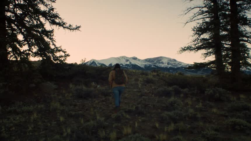 A solo hiker with a backpack walks up a grassy slope between trees with a view of a snow-covered mountain range at dusk