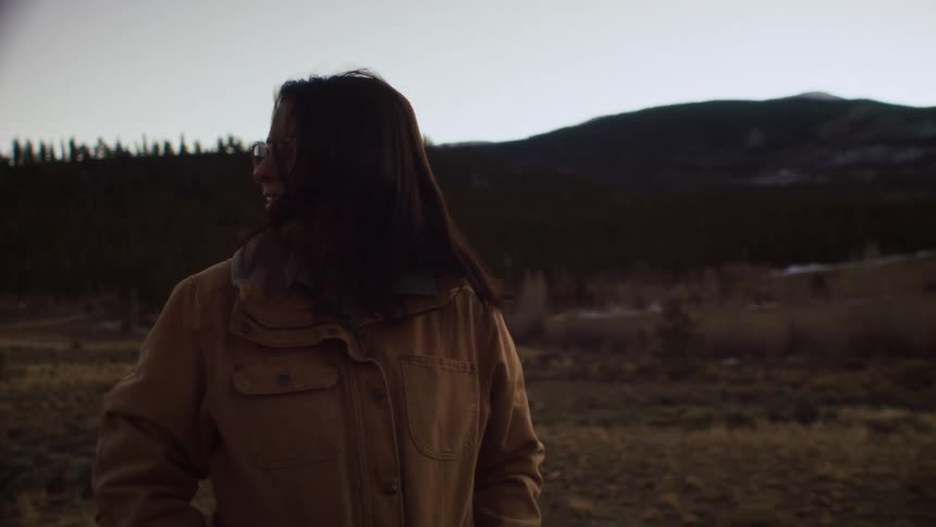 A woman in a rugged jacket stands in a windy mountain landscape, looking around with a gentle smile in slow motion