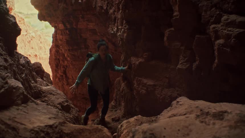 A female hiker with a backpack cautiously explores the inside of a dark and narrow red rock slot canyon in slow motion