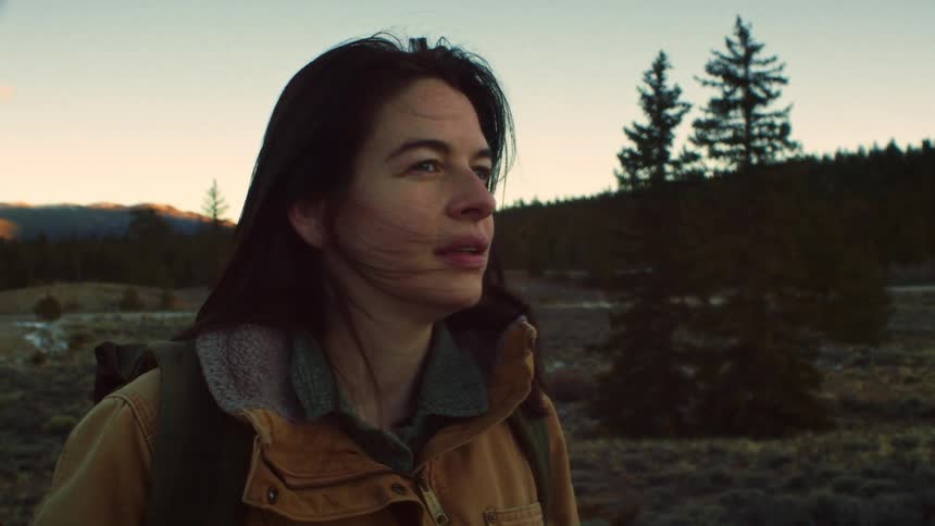 A contemplative female hiker with wind in her hair gazes out at the vast mountain landscape during a beautiful golden hour sunset