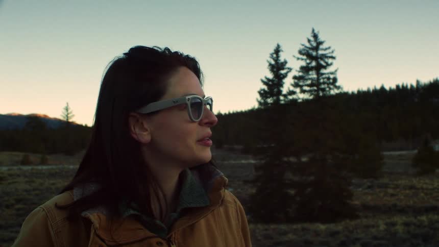 A young woman wearing sunglasses smiles as she looks out over a beautiful mountain landscape at dusk in slow motion