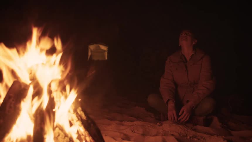 A woman relaxes by a roaring campfire in the sand at night, enjoying a moment of peace at her campsite