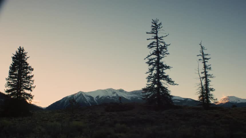 A lone hiker stands silhouetted on a hill, taking in the vast, snowy mountain landscape at sunrise