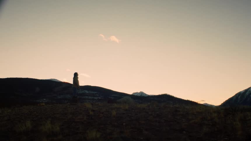 A slow dolly shot moves towards a lone person standing still on a hilltop, gazing out at the vast mountain range during a tranquil sunset