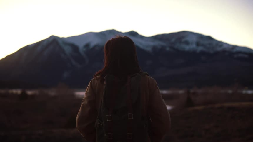 A female hiker with a backpack stands alone in a vast landscape, watching the sun rise over distant snow-capped mountains