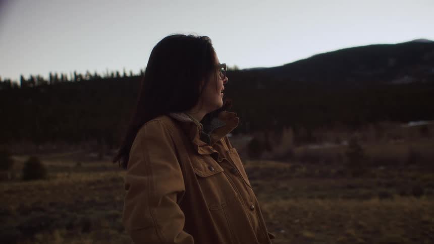 A woman in a brown jacket smiles peacefully while taking in the mountain view at dusk before walking out of frame