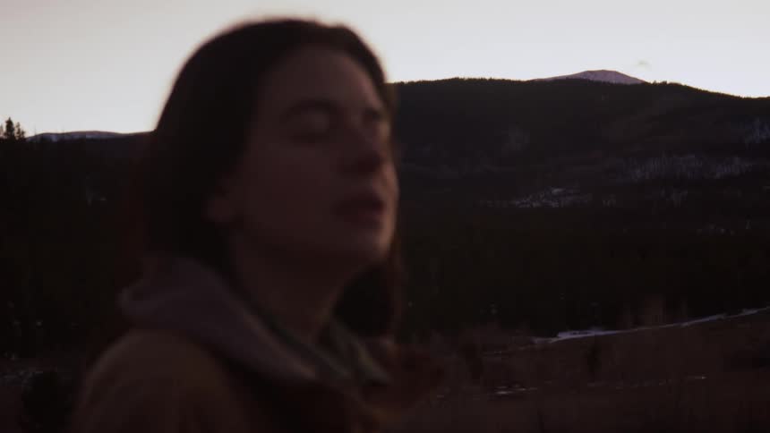 Close-up of a woman's peaceful face with eyes closed as the camera slowly racks focus to the mountain landscape behind her at dusk