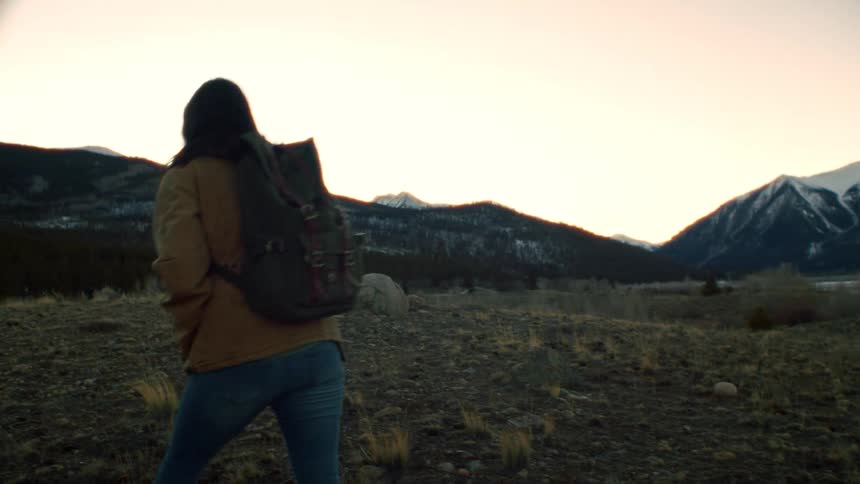 A woman with a backpack hikes away from the camera through a vast, windy mountain landscape in slow motion at sunset