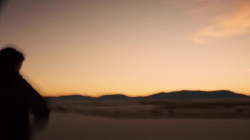 A shaky, blurry, handheld shot follows a woman walking through a vast white sand desert at sunset