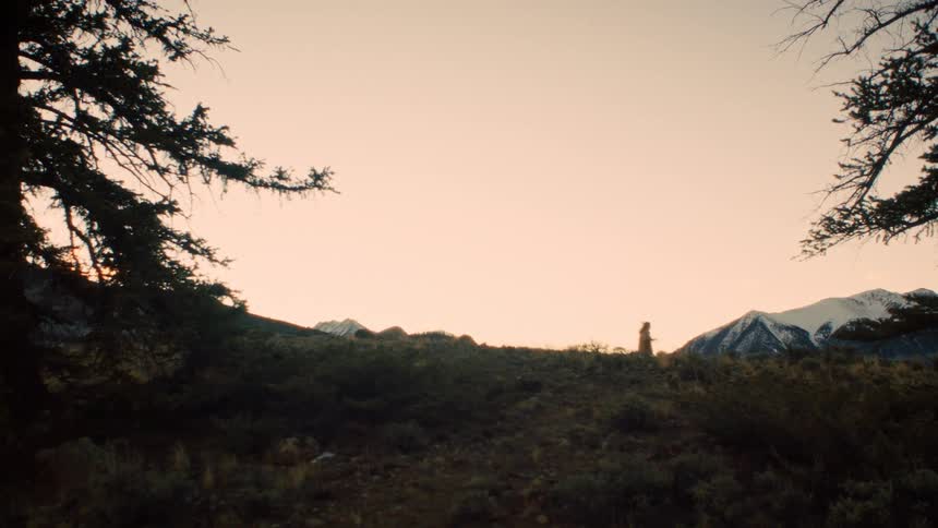 A silhouetted hiker walks along a mountain ridge with snow-capped peaks in the distance at dawn
