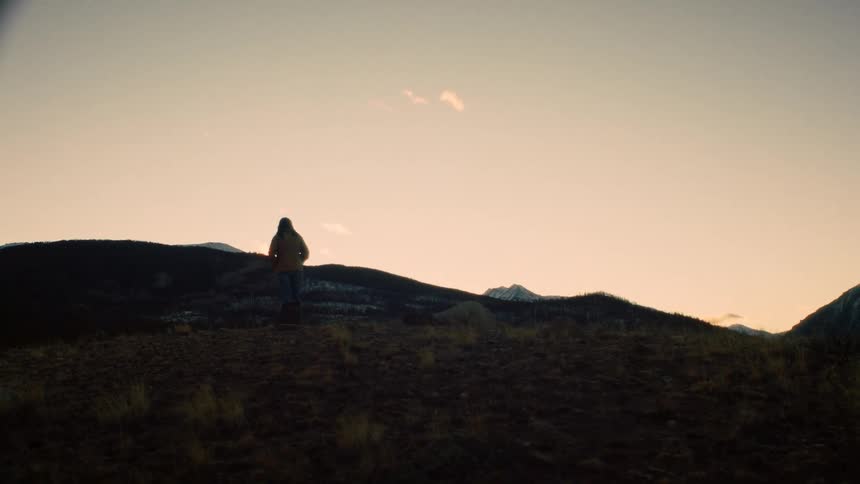 A silhouetted person stands on a remote hilltop during a hazy sunset, gazing at the distant mountain range