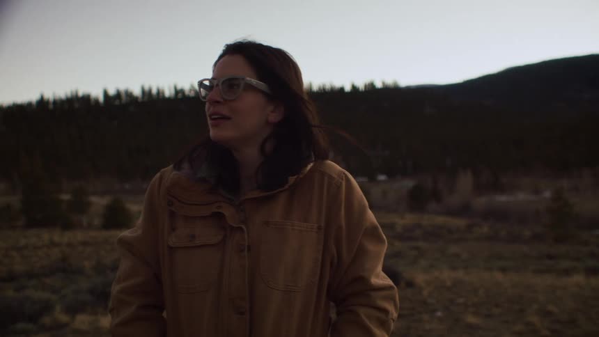 Slow motion shot of a woman in glasses and a brown jacket smiling as she looks out over a mountain landscape before turning and walking away at dusk