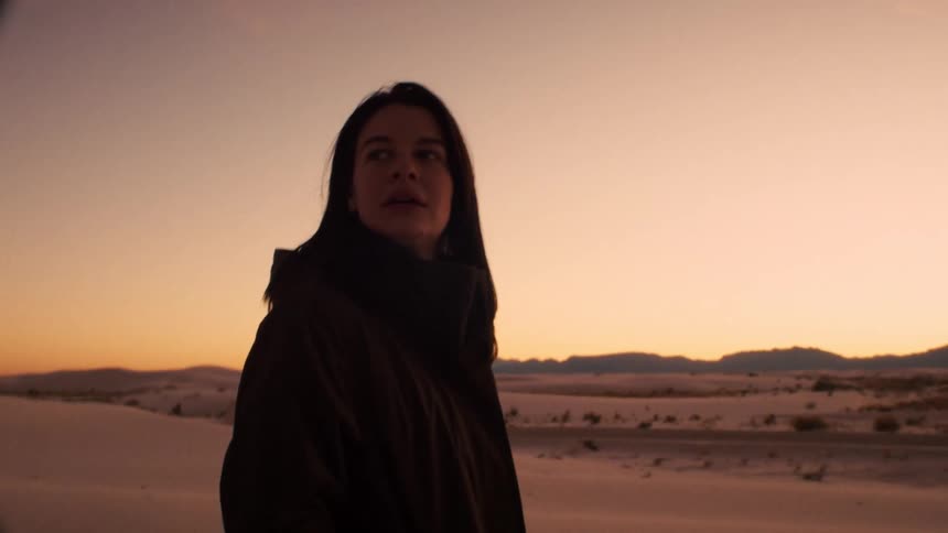 A contemplative woman stands amidst a vast white sand desert landscape, silhouetted by the warm glow of sunset