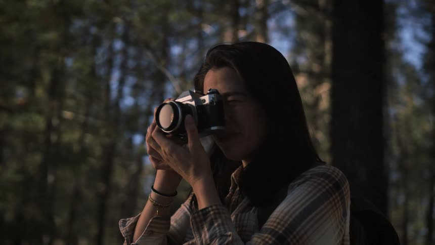 A smiling female photographer takes a picture with a vintage camera while exploring a beautiful sunlit forest