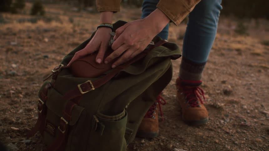A hiker takes a pair of eyeglasses from a leather case pulled from their rustic green backpack