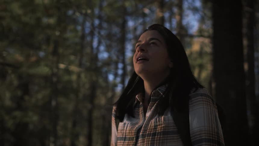 A woman with a backpack and a camera looks around in awe while hiking through a peaceful forest in slow motion