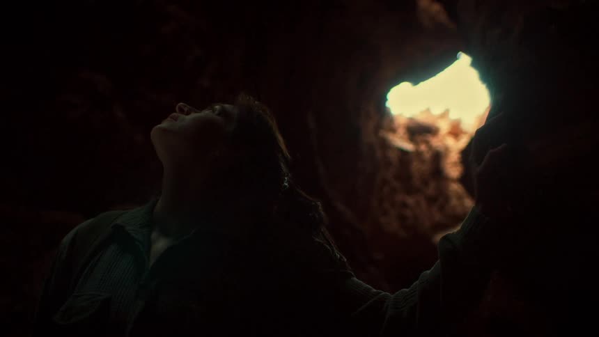A hopeful explorer smiles in a dark cave, gazing up at the light pouring through an opening above