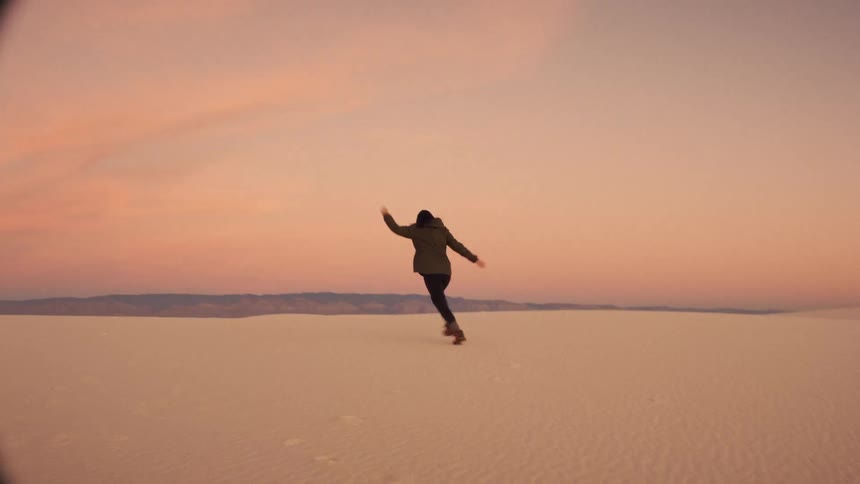 A woman playfully runs and twirls through a vast white sand desert during a beautiful sunset