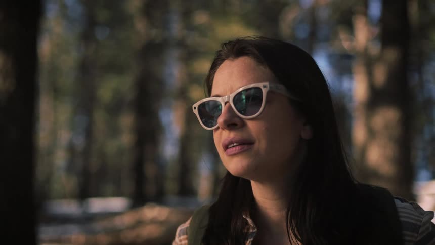 A woman wearing sunglasses looks up in wonder while hiking through a tranquil forest in slow motion