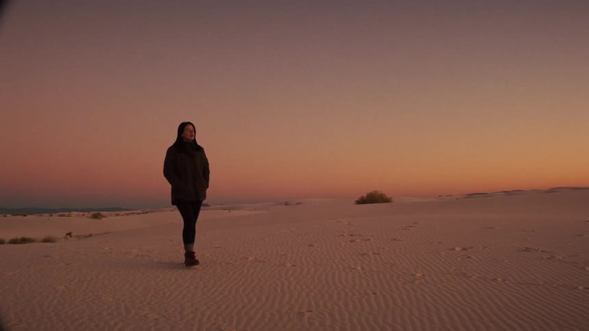 A woman stands and looks around in a vast, beautiful white sand desert during a vibrant sunset in slow motion