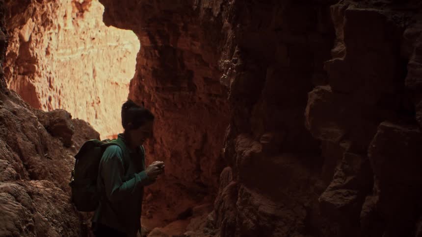 A female hiker in slow motion looks up with awe and wonder while exploring a dark, narrow slot canyon