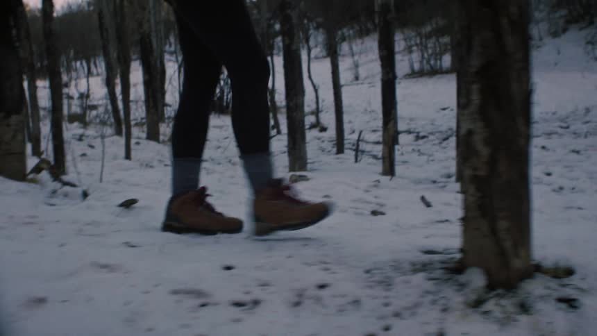 A low-angle slow-motion shot follows a hiker's legs and boots as they walk through a snowy forest