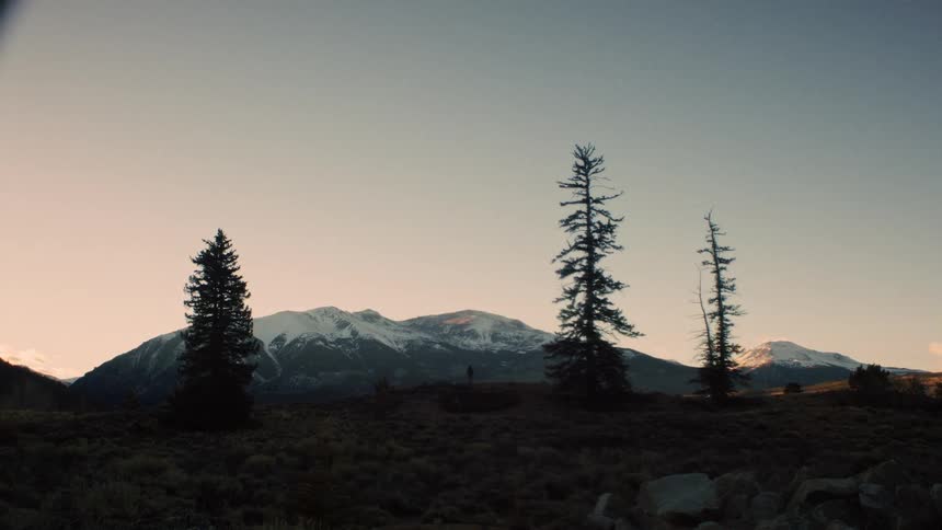 A silhouetted hiker walks along a ridge with pine trees against a stunning backdrop of snow-covered mountains at sunset