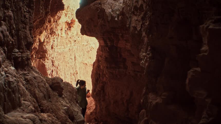 A woman with a backpack slowly explores the inside of a dark, narrow slot canyon, looking up in awe at the sunlit rock formations