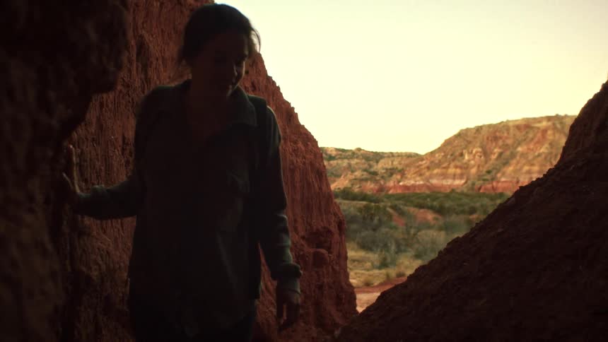 Slow motion shot of a woman with a backpack hiking towards the camera through a narrow, dark pass in a red rock canyon