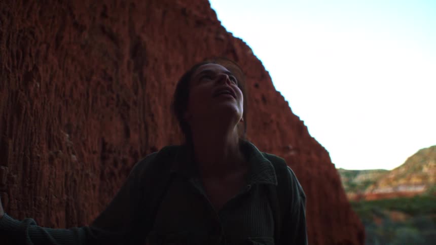A young female hiker in silhouette touches the textured red rock wall of a canyon cave and looks up in awe and wonder