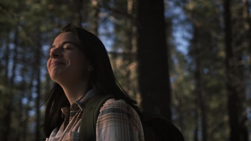 A peaceful female hiker closes her eyes and smiles, enjoying the warm sunlight filtering through the trees in a tranquil forest