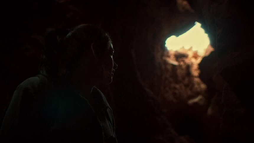 A silhouetted woman stands inside a dark cave, looking up towards a bright opening in the ceiling