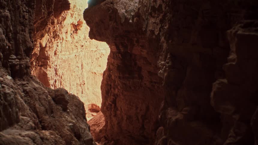 Looking out from a dark cave, a hiker carefully climbs through a sunlit, narrow canyon