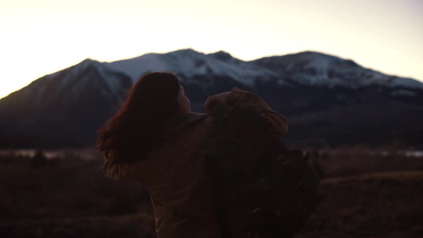 A female hiker puts on her backpack in slow motion while looking out at a stunning snow-capped mountain range at sunset