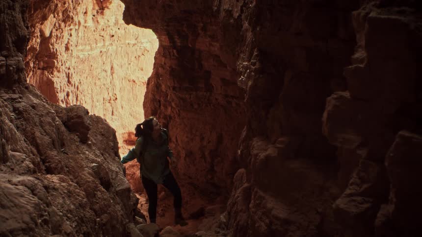 An adventurous woman with a backpack hikes through a dark and narrow slot canyon, cautiously navigating the rocky path towards the light