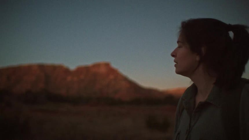 A female hiker gazes in awe at a red rock mountain landscape during a serene sunset, with the camera racking focus from the mountain to her face